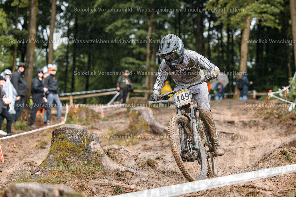 Mountainbike-Action im Wald | Ein Mountainbiker rast mit hoher Geschwindigkeit über einen anspruchsvollen, schlammigen Pfad in einem dichten Wald. Der Fahrer trägt einen schwarzen Helm und eine Schutzkleidung und trägt die Nummer 449. Im Hintergrund sind Zuschauer und weitere Bäume zu sehen. Der Pfad ist mit Wurzeln und Steinen übersät, was die Fahrt zusätzlich erschwert. - Realisiert mit Pictrs.com