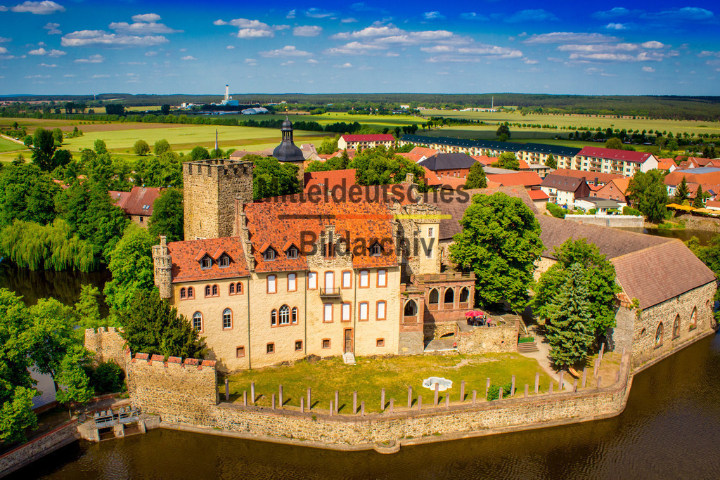 Wasserburg _ Schloss Flechtingen am See Börde-2386 | Dieses Luftbild zeigt die Wasserburg Flechtingen im Landkreis Börde in Sachsen-Anhalt an einem sonnigen Sommertag aus einer eindrucksvollen Vogelperspektive. Die historische Schloss- und Burganlage gehört zu den bekanntesten Wahrzeichen der Region und präsentiert sich in dieser Aufnahme von ihrer besonders farbenfrohen und malerischen Seite.Umgeben von den Wasserflächen des Schlosssees erhebt sich die Anlage mit ihren charakteristischen Türmen, Dächern und historischen Fassaden über die gepflegte Parklandschaft. Aus der Luft werden die harmonischen Proportionen des Ensembles und die besondere Lage am Wasser besonders deutlich sichtbar. Die Spiegelungen auf der Wasseroberfläche und die üppige Vegetation verleihen dem Motiv eine außergewöhnliche Atmosphäre.Das satte Grün der Bäume und Wiesen, die warmen Farbtöne der Gebäude und der strahlend blaue Himmel schaffen eine Bildwirkung, die an eine klassische Postkarte erinnert. Die Wasserburg Flechtingen zählt zu den schönsten historischen Anlagen Sachsen-Anhalts und ist ein beliebtes Ziel für Besucher, Fotografen und Erholungssuchende.Dieses Luftbild verbindet historische Architektur, Natur und sommerliche Farben zu einem stimmungsvollen Gesamtbild und zeigt die Wasserburg Flechtingen als eines der schönsten Ausflugsziele der Region Börde. - Realisiert mit Pictrs.com