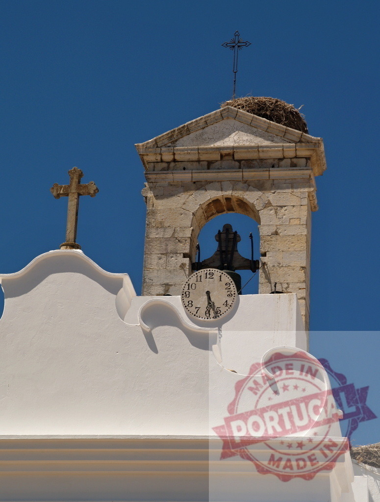Whitewashed church bell tower with stork nest and cross | Church bell tower features a cross, clock, and stork nest under a clear blue sky - Realisiert mit Pictrs.com