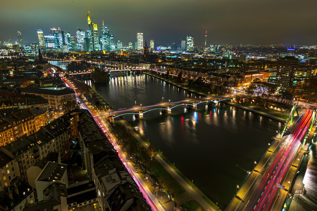 Wandbild - Nächtliches Frankfurt am Main: Eine Stadt der Lichter | Dieses beeindruckende Bild zeigt die nächtliche Skyline von Frankfurt am Main, einer der bedeutendsten Finanzmetropolen Europas. Die beleuchteten Wolkenkratzer spiegeln sich im ruhigen Wasser des Mains wider, während die beleuchteten Brücken, die den Fluss überspannen, die Stadtteile miteinander verbinden. Die langen Lichtspuren der fahrenden Autos auf den Straßen erzeugen ein dynamisches und lebendiges Bild. Die Mischung aus moderner Architektur und historischen Gebäuden verleiht der Szene eine besondere Atmosphäre. Die Farben und Lichter der Stadt strahlen eine einladende und aufregende Energie aus.