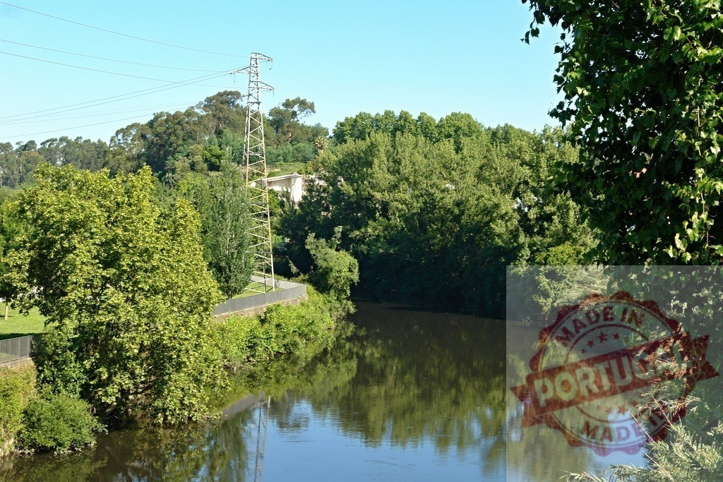 Electric power lines crossing tranquil river during summer day. Portugal - Norte | River reflecting a transmission tower and green trees under a clear sky - Realized with Pictrs.com