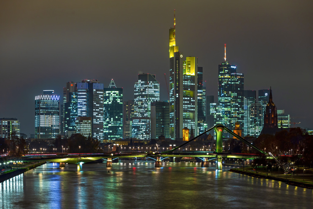 Wandbild - Frankfurts nächtlicher Glanz: Eine Skyline in Licht und Schatten | Dieses beeindruckende Bild zeigt die atemberaubende Skyline von Frankfurt am Main bei Nacht. Die imposanten Wolkenkratzer, darunter das markante Commerzbank-Hochhaus, ragen majestätisch in den Himmel und strahlen in einer Vielzahl von Farben und Lichtern. Die Beleuchtung der Gebäude spiegelt sich im ruhigen Wasser des Mains wider und verstärkt die Schönheit der Szene. Im Vordergrund sieht man die beleuchteten Brücken, die den Fluss überqueren und ein Gefühl der Verbindung zwischen den modernen Hochhäusern und den historischen Gebäuden vermitteln. Diese Aufnahme fängt die dynamische und pulsierende Atmosphäre einer der wichtigsten Finanzzentren Europas perfekt ein.