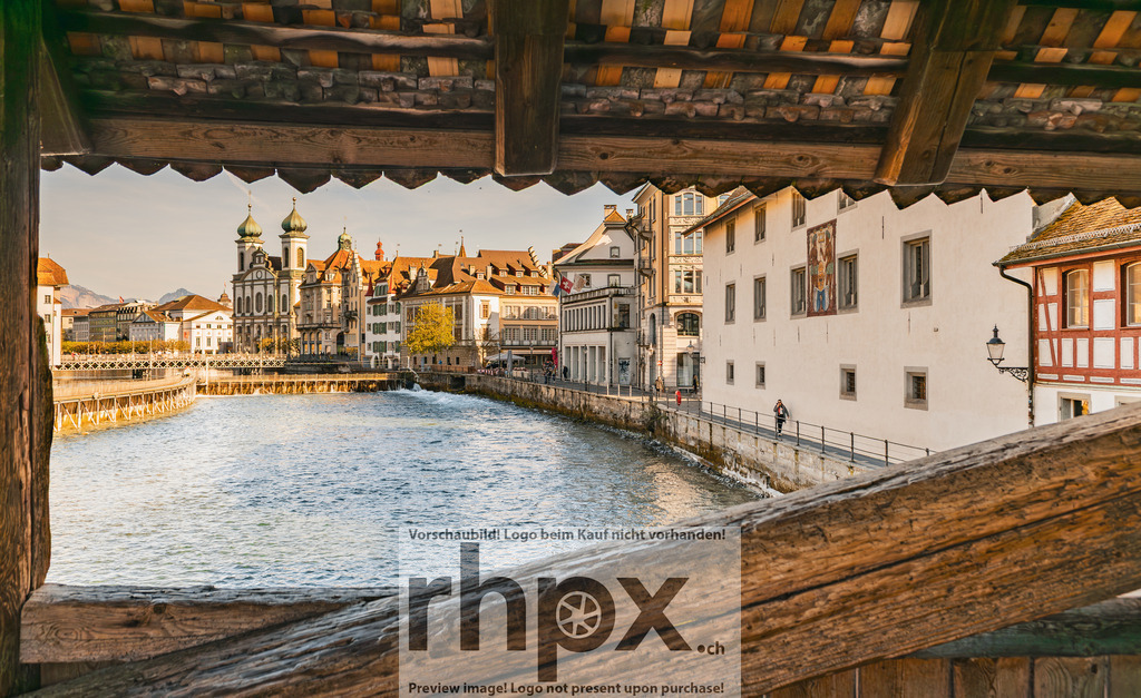Blick durch die Spreuerbrücke auf die Luzerner Altstadt | Gebälk. Fluss. Wahrzeichen.Durch das historische Holz der Spreuerbrücke öffnet sich der Blick auf die sonnenbeschienene Uferpromenade und die barocken Türme der Jesuitenkirche. Ein einzigartiges Fenster in die Geschichte Luzerns, wo Stadt und Wasser harmonisch verschmelzen.Wähle unter "Produktauswahl" dein Wunschformat: Vom klassischen Wandbild über Puzzle &amp; Tassen bis zum digitalen Download (z.B. als Handy-Hintergrund). - Realisiert mit Pictrs.com