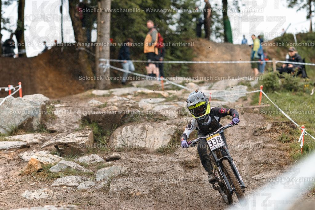 Downhill-Action im Wald | Das Bild zeigt einen Mountainbiker in voller Montur, der einen Downhill-Kurs durch einen bewaldeten Hang bewältigt. Der Fokus liegt auf dem Fahrer, der mit hoher Geschwindigkeit einen staubigen und steinigen Pfad entlang saust. Die Umgebung besteht aus Bäumen, Büschen und Felsen, die den Kurs säumen. Zuschauer beobachten das Rennen vom Rand aus. - Realisiert mit Pictrs.com