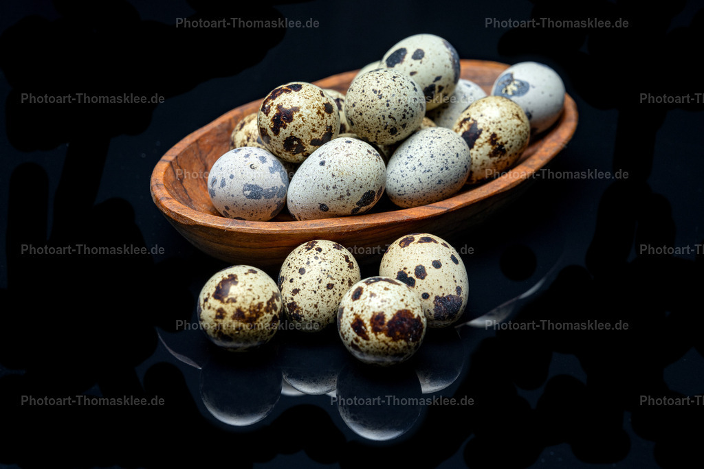 Gespiegelte Wachteleier - Elegantes Stillleben | Diese raffinierte Fine-Art-Fotografie inszeniert frische Wachteleier als elegantes Stillleben mit besonderem Twist. Die charakteristisch gesprenkelten Eier in ihrer natürlichen Farbpalette von Creme bis Graubraun ruhen in einer handgefertigten Olivenholzschale, deren warme, honigfarbene Maserung einen wunderschönen Kontrast zur kühlen Eleganz der schwarzen Spiegelplatte bildet. Das Spiegelbild der außerhalb der Schale platzierten Eier erzeugt eine faszinierende optische Illusion und verleiht der Komposition zusätzliche Tiefe. Die dramatische Beleuchtung betont die feinen Strukturen der Eierschalen und die lebendige Holzmaserung. Der fotografische Stil vereint klassische Stillleben-Ästhetik mit modernem Design und schafft eine zeitlose, fast meditative Atmosphäre, die den Betrachter zum Verweilen einlädt. - Realisiert mit Pictrs.com