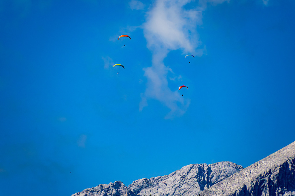 Tanz der bunten Schirme: Freiheit über den Gipfeln | Vor der imposanten Kulisse erhabener Felsmassive zieht eine Gruppe von Gleitschirmfliegern ihre Kreise am strahlend blauen Himmel. Zarte, luftige Wolkenbänder begleiten die farbenfrohen Schirme, die wie kleine Farbtupfer in der schier unendlichen Weite schweben. Das Bild fängt den magischen Moment absoluter Schwerelosigkeit und grenzenloser Freiheit ein – das stille Zusammenspiel aus menschlichem Abenteuergeist und der erhabenen Kraft der Natur. - Realisiert mit Pictrs.com