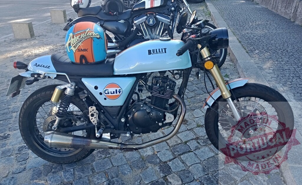 Bullit motorcycle parked on cobblestone street with Gulf logo and adventure motorcycle helmet | Stylish Bullit motorcycle with Gulf logo parked on a cobblestone street, featuring an adventure motorcycle helmet resting on the seat - Realisiert mit Pictrs.com