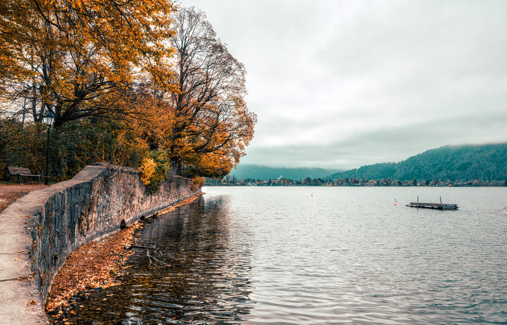 Uferblick über eine Kaimauer und herbstlich bunte Bäume auf den Tegernsee | Das Bild zeigt den Tegernsee im bayerischen Voralpenland im Herbst mit gelb gefärbten Bäumen am Ufer.Details zum Ort• Lage: Der See liegt im Landkreis Miesbach in Oberbayern, etwa eine Stunde südlich von München.• Landschaft: Umgeben von den bayerischen Alpen bietet die Region schöne Uferwege, Badestellen und traditionelle Orte wie Rottach-Egern oder Bad Wiessee. - Realisiert mit Pictrs.com