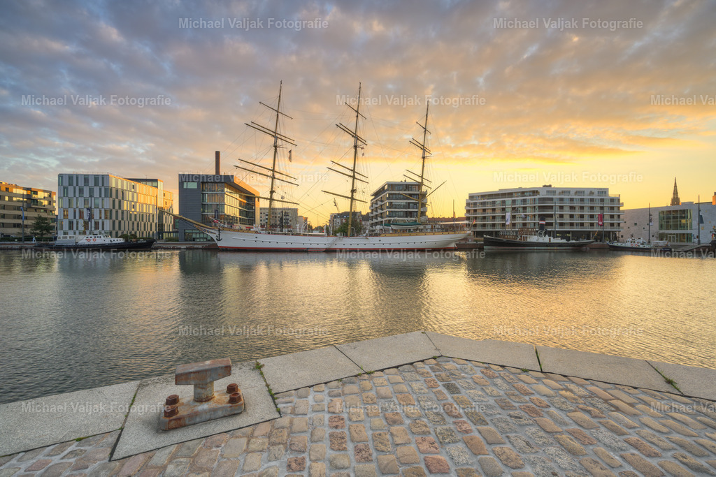 Schulschiff Deutschland im Neuen Hafen Bremerhaven | Im Zentrum des Neuen Hafens liegt das Schulschiff Deutschland – ein Dreimaster von 1927 und eines der letzten erhaltenen deutschen Segelschulschiffe. Fest vertäut an der Kaje, prägt es das Bild des historischen Hafenbeckens, das einst für die Auswanderung und den Überseehandel angelegt wurde. Heute ist das Schiff nicht nur technisches Kulturdenkmal, sondern auch ein lebendiger Teil des maritimen Erbes Bremerhavens – eingebettet in ein Umfeld aus Museumsschiffen, Promenade und Hafengeschichte. - Realisiert mit Pictrs.com
