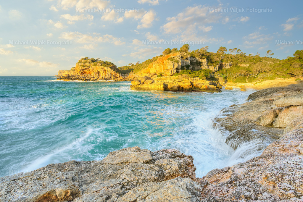 Cala s'Almunia auf Mallorca am frühen Morgen | Die Cala s’Almunia zeigt sich in einer klaren, warmen Morgenstimmung, in der die hellen Felsen der Bucht besonders deutlich hervortreten. Zwischen den kantigen Steinwänden bewegt sich das Wasser unruhig, läuft über die flachen Felsstufen und zieht sich wieder zurück, wodurch kurze, glänzende Spuren entstehen. Das Zusammenspiel aus warmen Felsfarben, schattigen Bereichen und dem lebendigen Meer verleiht diesem kleinen Küstenabschnitt eine direkte, natürliche Atmosphäre. - Realisiert mit Pictrs.com