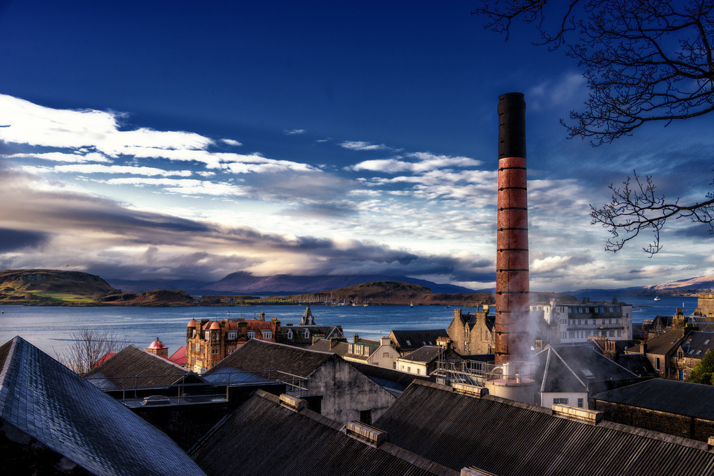 Oban Distillery | Ein hoher, roter Backsteinschornstein dominiert die Szenerie einer Küstenstadt, aus dem Wasserdampf aufsteigt. Im Hintergrund erstreckt sich eine weite Bucht mit bergiger Landschaft unter einem dramatischen Himmel mit Wolkenformationen. Die Komposition verbindet industrielle Architektur mit malerischer Natur und fängt das Licht eines Wintertages ein. - Realisiert mit Pictrs.com