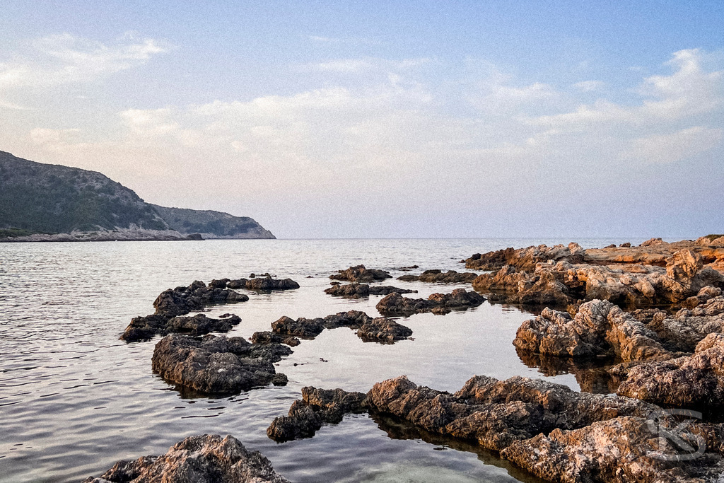 Abendruhe an der Cala Agulla – Exklusives Wandbild der mallorquinischen Felsenküste | Nahaufnahme von dunklen Küstenfelsen im ruhigen Wasser der Cala Agulla auf Mallorca während der Abenddämmerung mit Blick auf die entfernte Silhouette der Küstenlinie. - Realisiert mit Pictrs.com