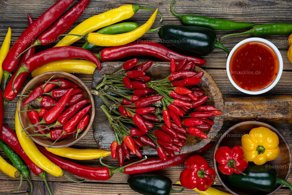 Farbenfrohe Gewürze: Chilischoten auf Holzuntergrund | Eine farbenfrohe Gewürzkomposition zeigt verschiedene Chili- und Paprikasorten auf rustikalem Holzuntergrund. Rote, gelbe und grüne Chilischoten verteilen sich harmonisch zwischen bunten Paprika. Eine Holzschale sammelt kleine rote Chilischoten, während ein rundes Holzbrett einen Strauß getrockneter Exemplare präsentiert. Eine weiße Schale enthält leuchtend rote Chilisauce. Die warmen Holztöne kontrastieren perfekt mit den lebendigen Gemüsefarben. Diese appetitliche Anordnung zeigt die kulinarische Vielfalt scharfer und milder Gewürze für kreative Kochexperimente. - Realisiert mit Pictrs.com
