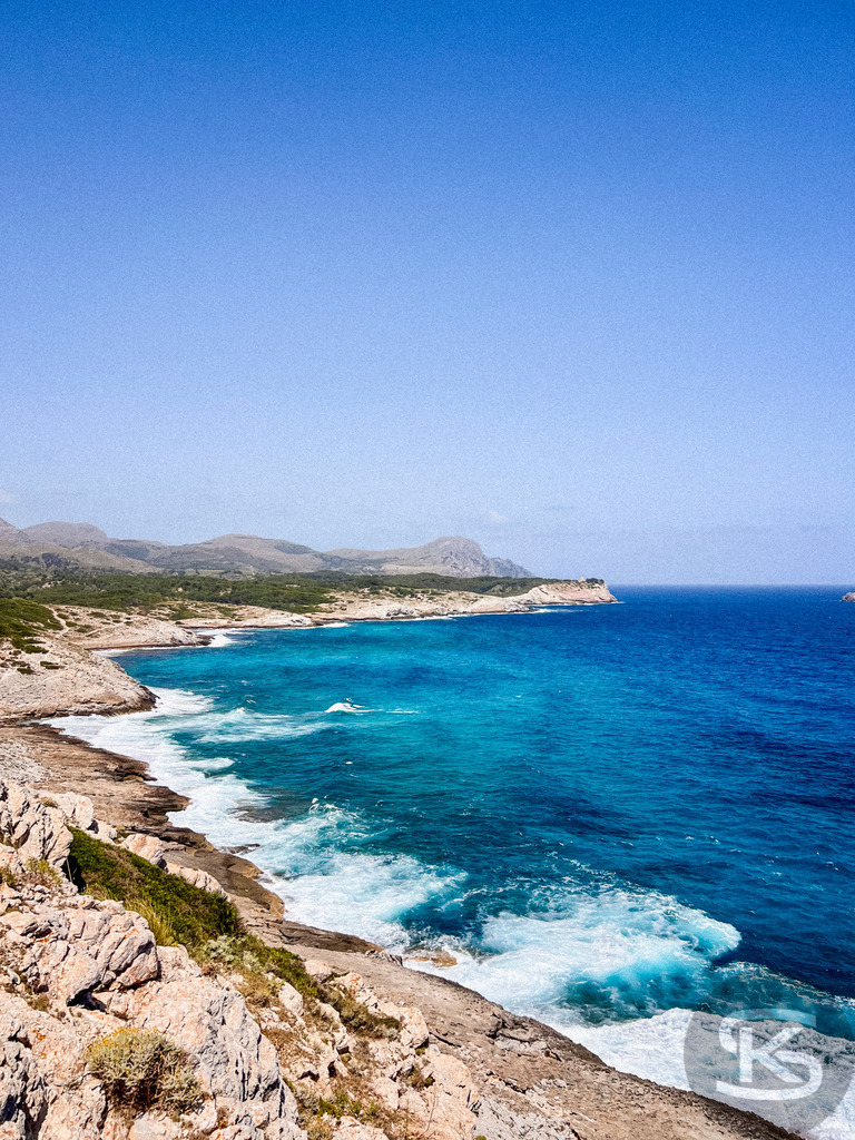 Spektakuläre Steilküste und Panoramablick bei Cala Mesquida, Mallorca | Panorama-Aufnahme einer felsigen Steilküste auf Mallorca bei Cala Mesquida mit schäumender Brandung am Ufer, tiefblauem Meer und einer weitläufigen Hügellandschaft unter klarem Himmel. - Realisiert mit Pictrs.com