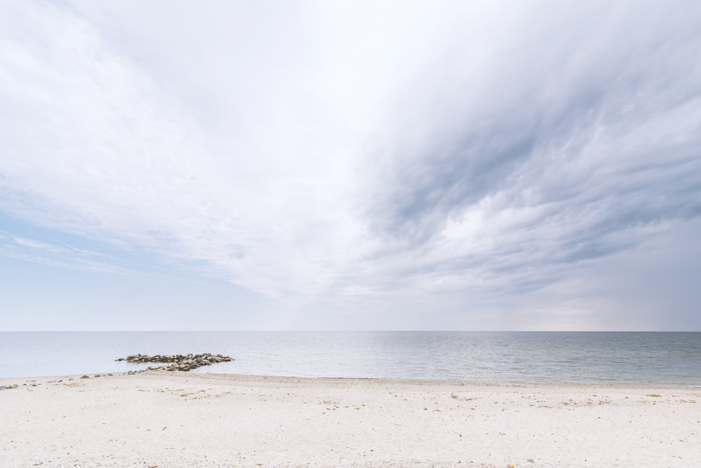 „Küstenruhe in sanften Nuancen – Minimalistisches FineArt‑Meeresbild | Dieses FineArt‑Wandbild zeigt eine ruhige Küstenszene in dezent reduzierten Farbtönen. Der helle Sand, die weiche Wasserlinie und der wolkenbetonte Himmel formen eine klare, ausgewogene Komposition. Eine schmale Steinmole setzt einen feinen, natürlichen Akzent, der die minimalistische Bildsprache unterstreicht, ohne die Ruhe des Motivs zu durchbrechen.Die zurückhaltende Farbpalette und die offene Weite verleihen dem Bild eine zeitlose Eleganz, die sich harmonisch in moderne, puristische Räume einfügt und eine Atmosphäre von Klarheit und Gelassenheit schafft.