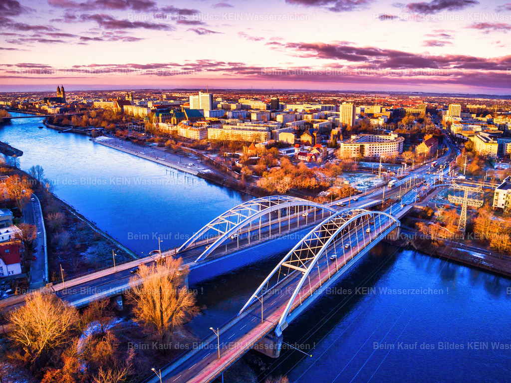 Magdeburg_Luftbild_jerusalembrücke_lila_Himmel_LR-0014 | Dieses Luftbild zeigt die beiden Jerusalembrücken in Magdeburg während der frühen Morgenstunden. Die markanten Brückenbauwerke überspannen die Elbe und verbinden die Innenstadt mit den östlichen Stadtgebieten. Aus der Vogelperspektive wird die elegante Linienführung der modernen Stahlkonstruktionen besonders deutlich sichtbar.Die Aufnahme lebt von ihrer außergewöhnlichen Lichtstimmung. Das Foto wurde farblich bewusst bearbeitet, wodurch intensive Lila-, Violett- und Magentatöne den Himmel und die Landschaft prägen. Die aufgehende Sonne sorgt für ein faszinierendes Farbenspiel, das den Brücken eine fast futuristische Wirkung verleiht und die Architektur eindrucksvoll hervorhebt.Im Hintergrund zeichnet sich die Silhouette der Magdeburger Innenstadt ab. Die Kombination aus moderner Infrastruktur, der Elblandschaft und dem spektakulären Morgenhimmel verleiht dem Bild eine besondere Dynamik. Gleichzeitig dokumentiert die Aufnahme die Bedeutung der Jerusalembrücken als wichtige Verkehrsverbindung und markantes Element der Stadtentwicklung.Dieses Luftbild verbindet technische Architektur mit kreativer Bildgestaltung und zeigt Magdeburg aus einer außergewöhnlichen und atmosphärischen Perspektive. - Realisiert mit Pictrs.com