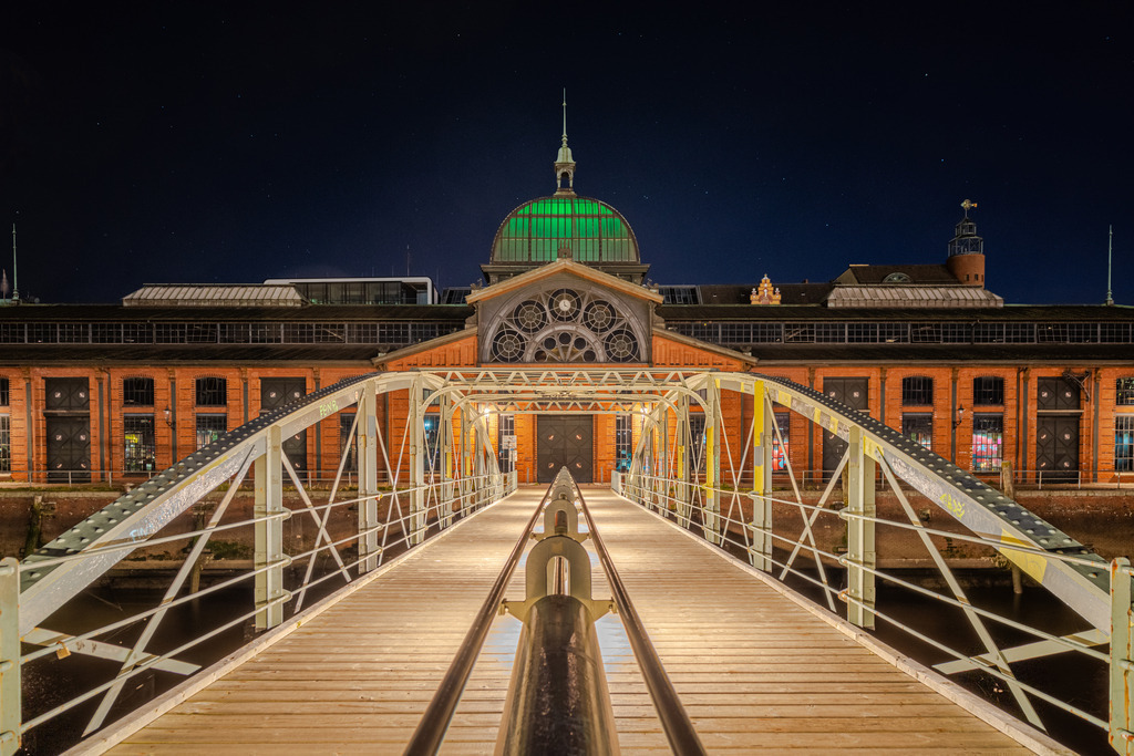 Alte Fischauktionshalle | Das Bild zeigt die beeindruckende alte Fischauktionshalle in Hamburg bei Nacht. Im Vordergrund führt eine beleuchtete Brücke direkt auf den Haupteingang der historischen Halle zu. Die rote Backsteinfassade der Halle ist durch zahlreiche Fenster unterteilt und wird durch eine große, kunstvolle Fensterrosette über dem Eingang verziert. Die beleuchtete Kuppel der Fischhalle strahlt in einem grünen Licht, was dem Gebäude ein majestätisches Aussehen verleiht. Die gesamte Szene wirkt ruhig und klar, der Himmel ist dunkel und die präzise Beleuchtung hebt die architektonischen Details der Fischauktionshalle hervor.