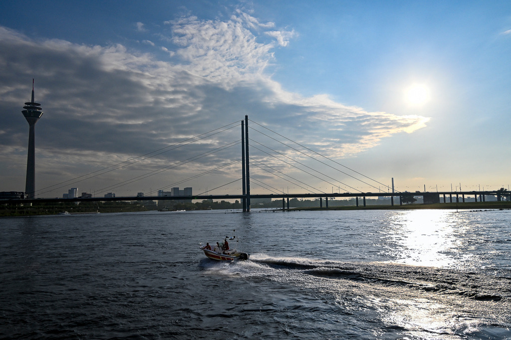 DLRG im Einsatz vor der Düsseldorfer Skyline | In diesem Foto sieht man ein Boot der Deutschen Lebens-Rettungs-Gesellschaft (DLRG), das mit hoher Geschwindigkeit über den Rhein fährt und eine Gischtwelle hinter sich herzieht. Im Hintergrund erstreckt sich die imposante Rheinkniebrücke über den Fluss. Links ragt der Fernsehturm Düsseldorf empor, während die Sonne am rechten Bildrand hell leuchtet und das Wasser glitzern lässt. Die Szene vermittelt ein Gefühl von Dynamik und Einsatzbereitschaft vor der Kulisse der Landeshauptstadt Nordrhein-Westfalens. - Realisiert mit Pictrs.com