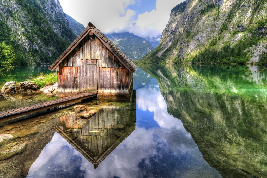 Wandbild - Ruhiges Spiegelbild: Hütte am Bergsee | Dieses atemberaubende Bild zeigt eine rustikale Holzhütte, die am Rande eines kristallklaren Bergsees steht. Die Hütte spiegelt sich perfekt im ruhigen Wasser, das von den umgebenden Bergen und dem blauen Himmel umrahmt wird. Die Szene ist idyllisch und friedlich, mit üppig grünen Wäldern, die sich in den stillen See erstrecken, und majestätischen Bergen, die in der Ferne aufragen. Ein schmaler Holzsteg führt zur Hütte, die Einsamkeit und Ruhe inmitten der Natur ausstrahlt. Der Himmel ist teilweise bewölkt und reflektiert sanft im Wasser, was der gesamten Komposition eine harmonische und beruhigende Wirkung verleiht.