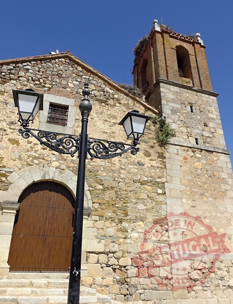 Stone church with bell tower and stork nest. Spain - Extremadura | Historic stone church featuring a bell tower, stork nest, and decorative street lamp - Realisiert mit Pictrs.com