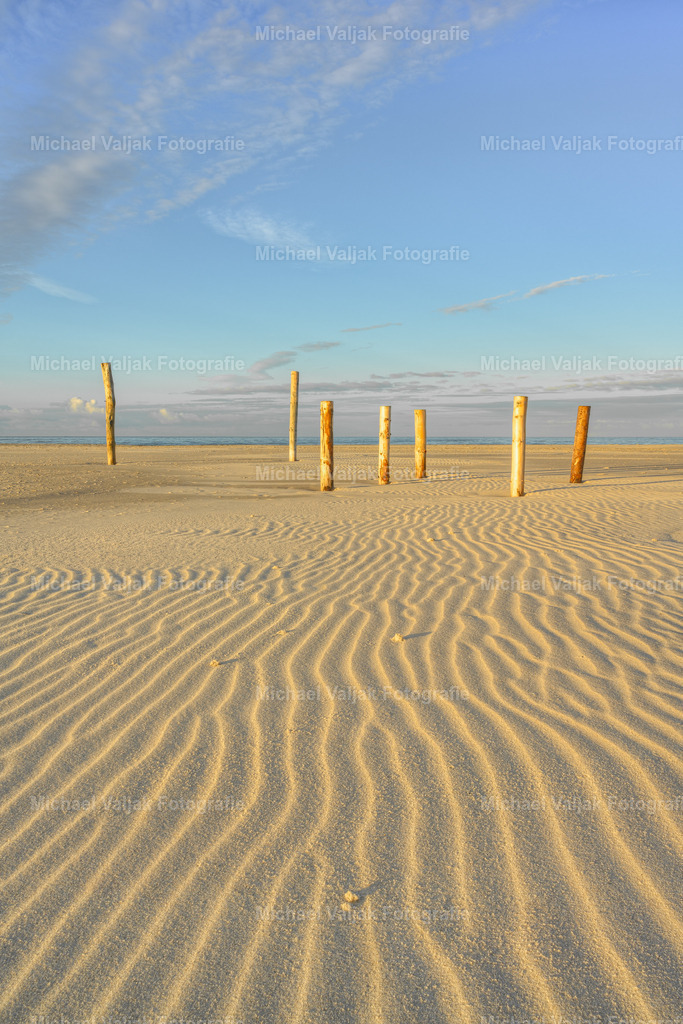Holzpfähle auf dem Kniepsand auf Amrum | Der Kniepsand auf Amrum ist eine faszinierende geologische Erscheinung. Obwohl er scheinbar ein Teil der Insel ist, gehört er geologisch gesehen nicht zu Amrum und ist eine eigenständige Sandbank. Diese Sandbank bietet einen 15 Kilometer langen und bis zu 1,5 Kilometer breiten Sandstrand, der für lange, einsame Strandwanderungen einlädt. Der Kniepsand dient auch als natürlicher Schutz für die Insel gegen Sturmfluten und orkanartige Wetterbedingungen. Die Pfähle, die man im Sand sieht, sind Zeugen menschlicher Aktivitäten und tragen zur einzigartigen Atmosphäre dieses Ortes bei. - Realisiert mit Pictrs.com