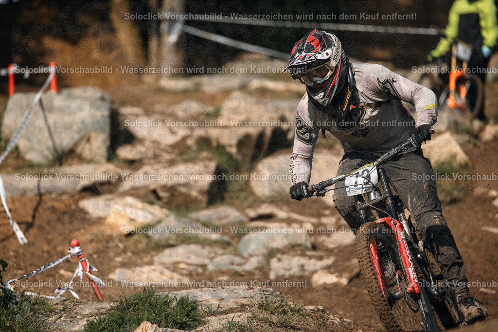 Mountainbiker im Sturzflug | Ein Mountainbiker stürzt sich mit hoher Geschwindigkeit einen steilen, steinigen Pfad hinunter. Er trägt einen schwarzen Helm und Schutzkleidung und fährt ein rotes Mountainbike mit schwarzem Rahmen. Der Hintergrund zeigt eine felsige Landschaft mit Bäumen und Büschen. - Realisiert mit Pictrs.com