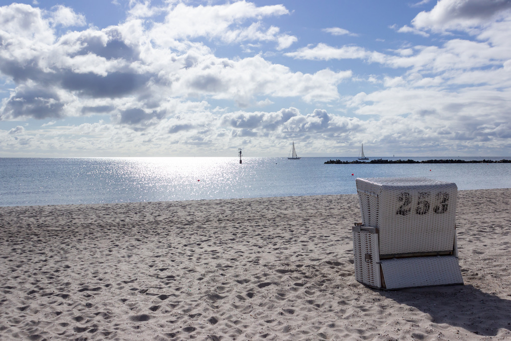 Wandbild: Strandkorb am Strand in Damp | Dieses Wandbild im Querformat zeigt einen Strandkorb an der Ostsee in Damp. Am blauen Himmel sind viele Wolken zu sehen. In der Ferne sind zwei Segelboote zu sehen.   - Realisiert mit Pictrs.com