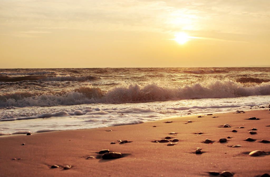Wandbild: Sonnenaufgang an der Ostsee | Dieses Wandbild im Querformat zeigt einen malerischen Sonnenaufgang an der Ostsee. Die Sonne spiegelt sich auf dem Meer. Im Vordergrund ist der schöne Sandstrand zu sehen, an dem Steine liegen. Der Himmel leuchtet in einem schönen orange.  - Realisiert mit Pictrs.com
