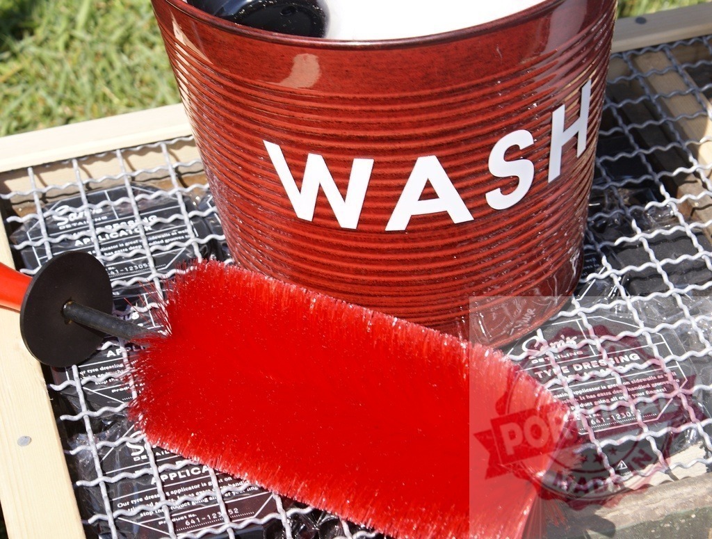 Red wash bucket with detailing brush for cleaning | Red metal bucket with WASH text next to a red wheel cleaning brush on a metal grid - Realisiert mit Pictrs.com