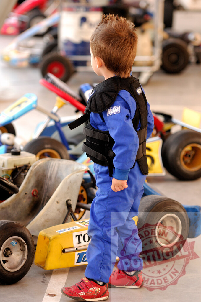 Young boy wearing protective clothing standing near go-karts | Little boy wearing a blue racing suit and a black protective vest standing near go-karts in a kart racing circuit - Realisiert mit Pictrs.com