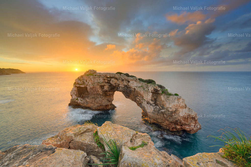 Goldenes Erwachen über Es Pontàs auf Mallorca | Die Sonne steigt langsam über den Horizont und taucht Es Pontàs in ein warmes Leuchten. Der Felsbogen hebt sich klar vor dem farbintensiven Himmel ab, während das Meer die ersten Strahlen glitzernd zurückwirft. Ein stiller Moment, in dem der Tag sichtbar Gestalt annimmt. - Realisiert mit Pictrs.com