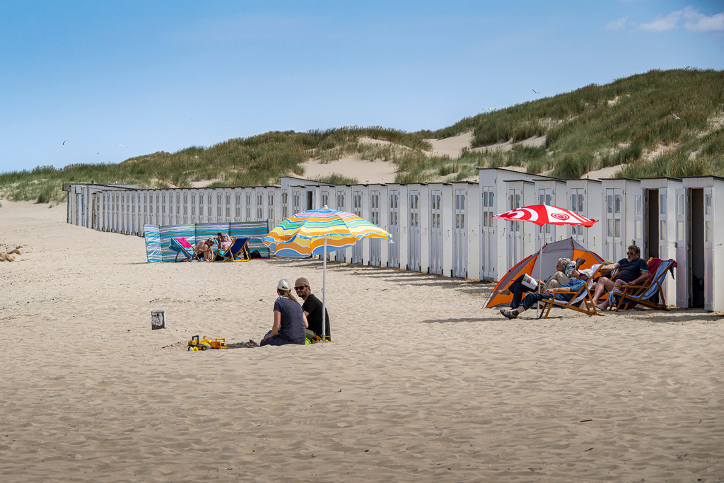 Sommerliche Strandidylle auf Texel | Ein unbeschwerter Ferientag am breiten Sandstrand der Nordseeinsel Texel: Vor der charakteristischen Reihe weißer Strandhäuschen genießen Urlauber das sonnige Wetter. Bunte Sonnenschirme, Windschutze und Liegestühle setzen fröhliche Farbakzente im feinen Sand. Während im Vordergrund ein Paar im Schatten verweilt, entspannen andere Gäste geschützt vor den Dünen. Die sanften Grashügel im Hintergrund und der weite Himmel vermitteln ein Gefühl von unendlicher Freiheit, Ruhe und klassischer Nordsee-Gemütlichkeit. - Realisiert mit Pictrs.com