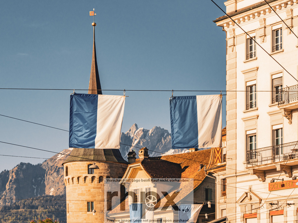 Luzern mit Pilatus und Fahnen im Morgenlicht | Luzern im klaren Morgenlicht, eingerahmt von zwei blau-weissen Fahnen. Dahinter zeichnet sich der Pilatus scharf vor dem Himmel ab.  - Realisiert mit Pictrs.com