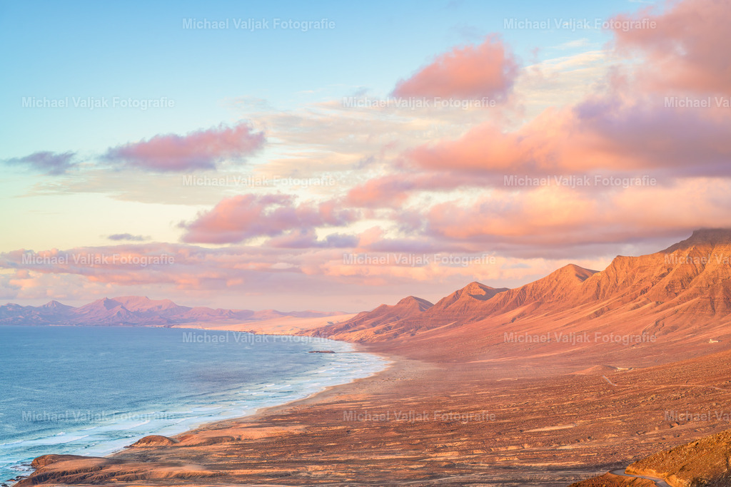 Mirador de Cofete auf Fuerteventura | Vom Mirador de Cofete fällt der Blick weit über die endlose Westküste Fuerteventuras, die in der warmen Abendsonne in weichen Gold‑ und Orangetönen leuchtet. Die langen, geschwungenen Linien der Berge ziehen sich bis hinunter zum Strand, der wie ein heller Streifen zwischen Fels und Meer liegt. Das Licht betont die Formen der Landschaft und lässt die Schatten langsam länger werden, während der Atlantik in tiefem Blau schimmert. Ein Moment, in dem die Weite, die Stille und die besondere Magie dieser Aussicht spürbar werden. - Realisiert mit Pictrs.com