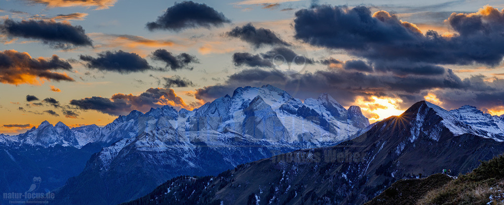 R6NF7212_20251005-Pano | Eine atemberaubende Panoramaaufnahme der schneebedeckten Dolomiten bei Sonnenuntergang oder Sonnenaufgang. Im Zentrum dominiert die majestätische Marmolada mit ihrem Gletscher, umgeben von weiteren imposanten Gipfeln. Die tief stehende Sonne taucht den Himmel in warme Gold- und Orangetöne, die sich mit den dunklen, dramatischen Wolken mischen. Die unteren Berghänge zeigen noch Spuren von Vegetation, während die höheren Lagen bereits von frischem Schnee bedeckt sind, was auf den Übergang zwischen Herbst und Winter hindeutet. Ein kleiner Lichtstrahl bricht hinter einem Berg auf der rechten Seite hervor und beleuchtet die Szenerie. - Realisiert mit Pictrs.com
