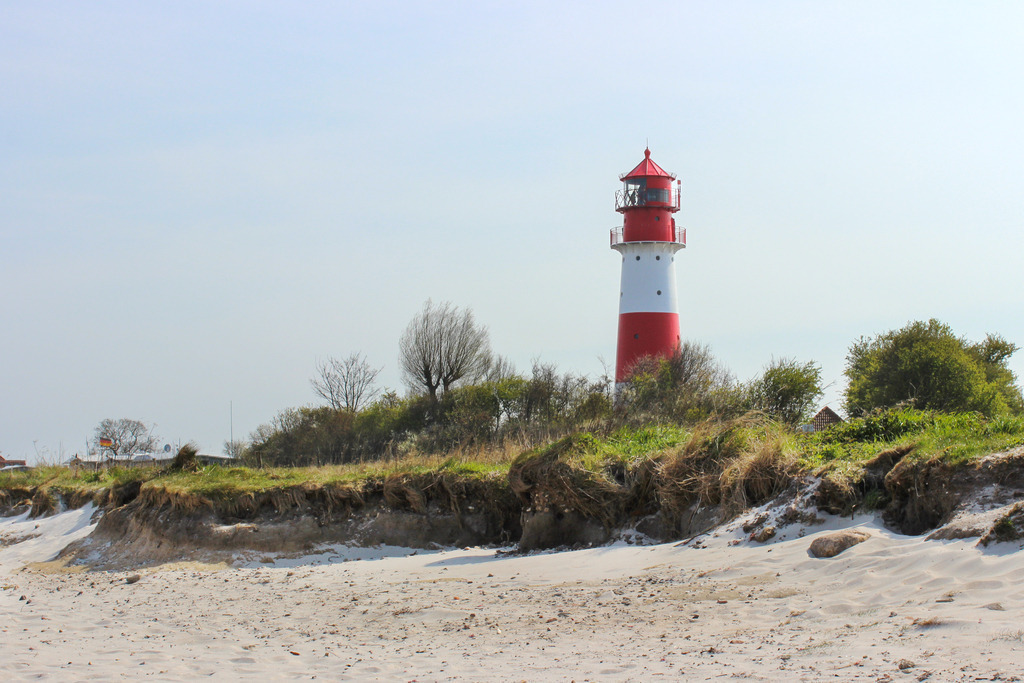 Wandbild: Leuchtturm in Falshöft im Frühling | Dieses Wandbild im Querformat zeigt den Strand und den Leuchtturm in Falshöft im Frühling. Im Vordergrund ist der Strand zu sehen. Der Leuchtturm steht inmitten von Gräsern und Büschen. Der blaue Himmel ist fast wolkenlos.  - Realisiert mit Pictrs.com