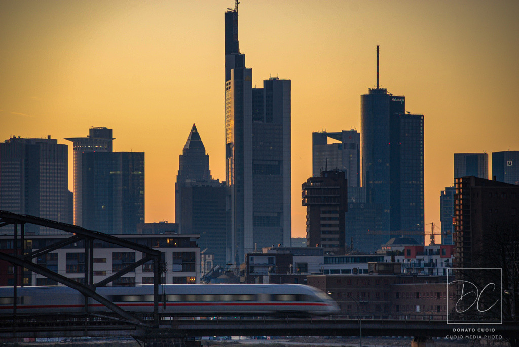 Frankfurt Skyline Foto: ICE auf Deutschherrnbrücke bei Sonnenuntergang | Dieses stimmungsvolle Foto fängt den Zauber eines Sonnenuntergangs über der pulsierenden Mainmetropole Frankfurt ein. Im Vordergrund überquert ein moderner ICE-Zug elegant die historische Deutschherrnbrücke, ein Symbol für Bewegung und Fortschritt. Im Hintergrund erhebt sich die beeindruckende Frankfurter Skyline mit ihren markanten Wolkenkratzern, die in warmes, goldenes Licht getaucht sind. Die Aufnahme, entstanden im Februar 2008, zeigt die Stadt in einem Moment der Ruhe und Schönheit, eingefangen in einem perfekten Zusammenspiel von Architektur, Natur und Technologie. - Realisiert mit Pictrs.com