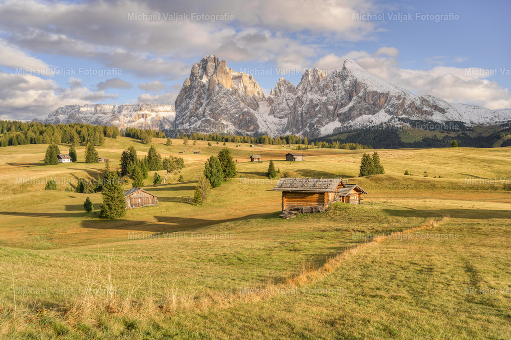 Seiser Alm im Herbst | Blick über die Almwiesen und Almhütten der Seiser Alm in Südtirol zu den bereits mit Schnee bedeckten Bergen Langkofel und Plattkofel. Die Sonne taucht die Landschaft in ein warmes Licht. - Realisiert mit Pictrs.com