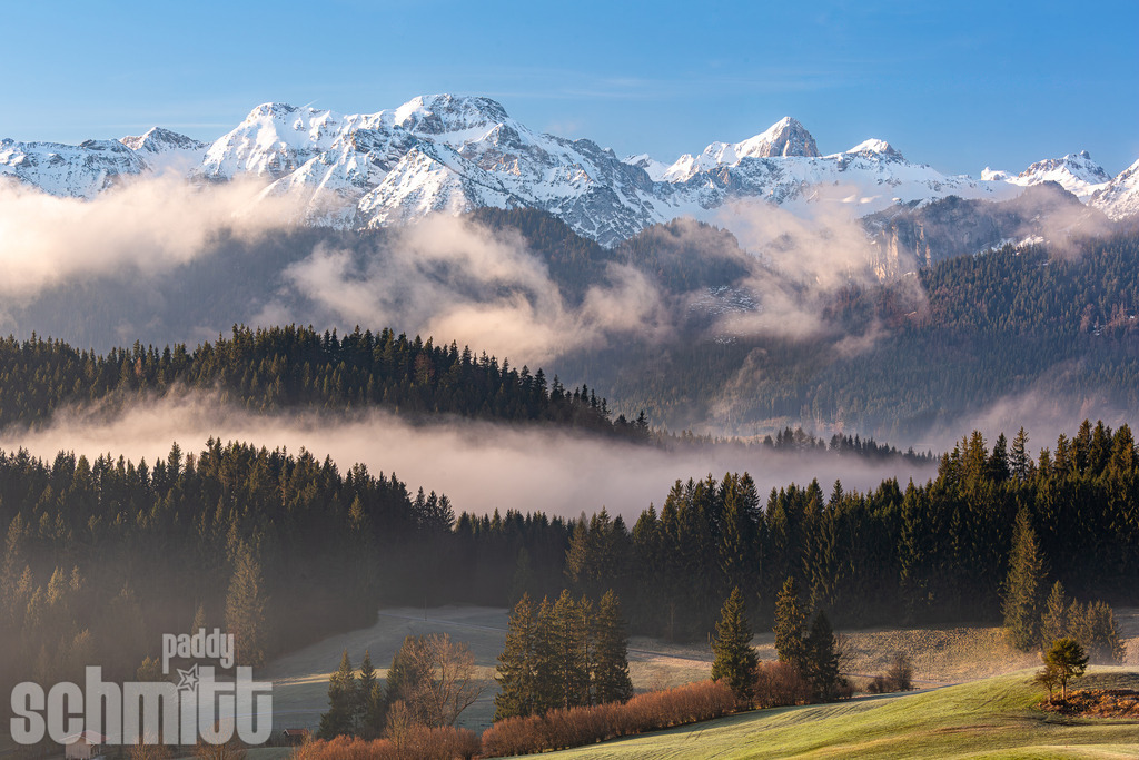 Morgennebel vor verschneiter Bergkulisse im Frühling | Impulsiver Morgennebel liegt sanft über der Frühlingslandschaft, während sich dahinter die noch verschneiten Berge erheben. Eine ruhige und stimmungsvolle Aufnahme vom frühen Morgen im Allgäu. - Realisiert mit Pictrs.com