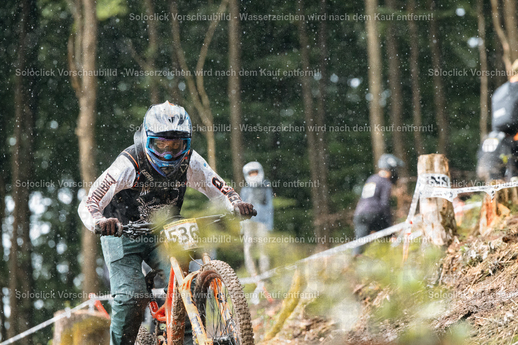 Mountainbike-Abenteuer im Regen | Das Bild zeigt einen Mountainbiker in voller Ausrüstung, der einen steilen, bewaldeten Hang hinunterfährt. Der Fahrer trägt einen Helm, eine Brille, Handschuhe und spezielle Kleidung. Er trägt die Nummer 536. Der Hintergrund besteht aus dichtem Grün und Bäumen. Es regnet, was die Szene dynamisch und herausfordernd erscheinen lässt. - Realisiert mit Pictrs.com