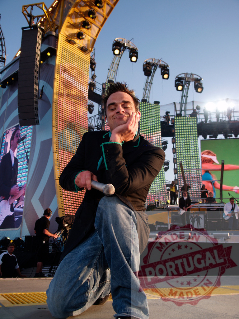 Robbie Williams kneeling on stage holding microphone | Robbie Williams kneeling on stage during a concert, holding a microphone and smiling - Realized with Pictrs.com