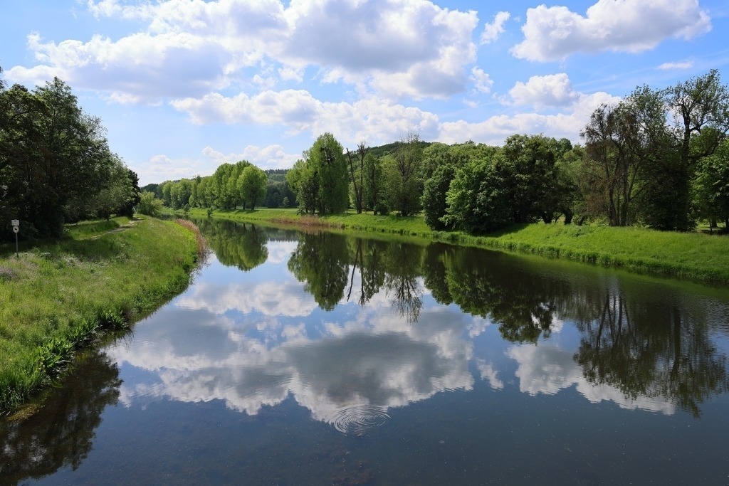 Wolkenspiegelung im Fluss der Weißen Elster | Die Aufnahme zeigt die ruhige Oberfläche der Weißen Elster am Wehr von Bad Köstritz, in der sich ein dramatischer Himmel mit großen, weichen Wolken spiegelt. Durch das fast unbewegte Wasser wirkt die Spiegelung beinahe wie ein zweites Bild unterhalb des Horizonts. Die Ufer des Flusses rahmen die Szene natürlich ein – mit Bäumen und Wiesen im Hintergrund – und verleihen dem Motiv Tiefe.Das Licht ist sanft und diffus. Dadurch entstehen feine Kontraste zwischen Himmel, Wasser und Landschaft. Die Wolken wirken plastisch und ziehen den Blick in die Spiegelung hinein, sodass Realität und Reflexion fast miteinander verschmelzen.Die Stimmung des Fotos ist still, meditativ und naturverbunden. Besonders die Kombination aus Himmel und Wasser vermittelt Weite und Gelassenheit. - Realisiert mit Pictrs.com