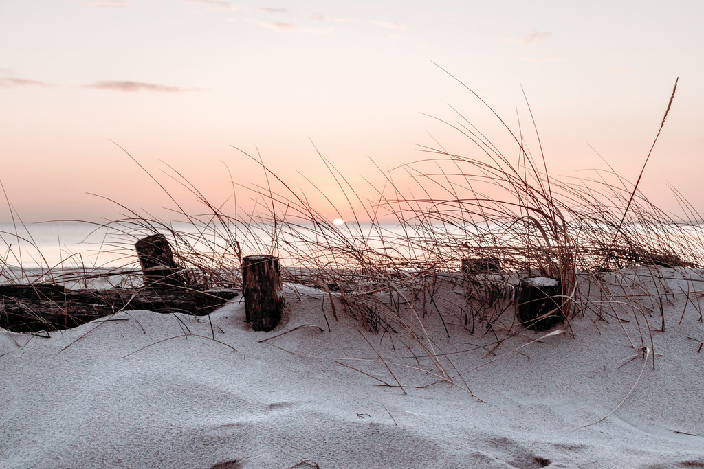 Wandbild: Düne und Strandhafer im Sonnenaufgang | Dieses Wandbild im Querformat zeigt eine kleine Düne bewachsen mit Strandhafer am Meer. Einige kleine Holzpfähle sorgen für einen schönen Blickfang. Im Hintergrund ist die aufgehende Sonne über dem Meer zu sehen, die den Himmel in ein wunderschönes pastellartiges rotorange taucht. Der Strandsand bringt einen natürlichen Sandton ins Bild und das warme Rot erzeugt eine wohnliche Stimmung. Holen Sie sich diesen wunderschönen Sonnenaufgang am Meer auf Leinwand, Aluminium-Platte oder Acrylglas. Ideal fürs Wohnzimmer, Schlafzimmer, Küche, den Arbeitsplatz oder die Ferienwohnung.   - Realisiert mit Pictrs.com