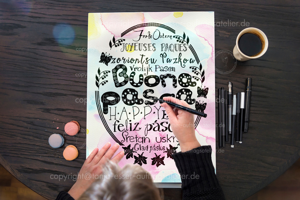 Hand lettering - A woman sits at the table and draws "Happy Easter" as text in several languages ​​by hand | Hand Lettering - Eine Frau sitzt am Tisch und zeichnet den Text Frohe Ostern auf Papier
. Das Papier ist mit Aquarell Klecksen in Pastellfarben bemalt. Von oben fotografiert.