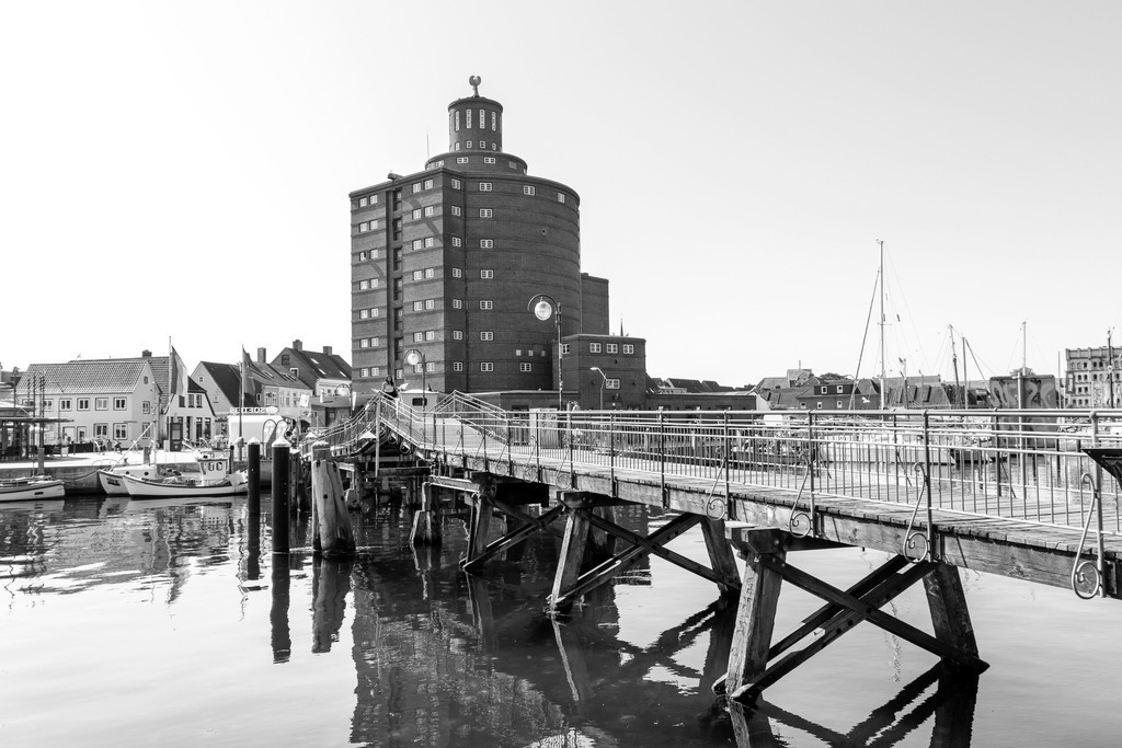 Wandbild: Rundsilo am Hafen in Eckernförde in Schwarz-Weiß | Dieses Wandbild im Querformat zeigt das Rundsilo und die Fußgängerbrücke am Hafen in Eckernförde in Schwarz-Weiß. Auf dem Wasser ist eine schöne Spiegelung zu sehen. Der blaue Himmel ist fast wolkenlos.  - Realisiert mit Pictrs.com