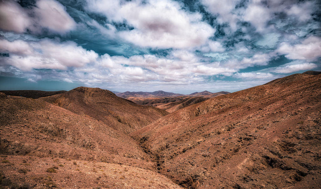 Blick in den felsigen Barranco de las Peñitas, Fuerteventura | Das Bild zeigt den Aussichtspunkt Mirador de las Peñitas auf der Insel Fuerteventura.Lage: Er befindet sich in der Gemeinde Betancuria.Aussicht: Der Punkt bietet einen Blick auf das Barranco de las Peñitas, ein tiefes Tal mit einer kleinen Oase.Umgebung: In der Nähe liegt der Stausee Embalse de las Peñitas und die Ermita de las Peñitas.Wandern: Die Gegend ist beliebt für Wanderungen durch die felsige Landschaft, beispielsweise zum Arco de las Peñitas. - Realisiert mit Pictrs.com