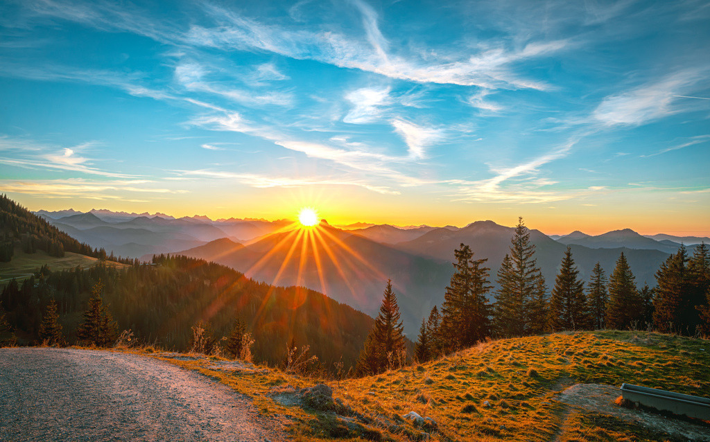 Herbstlicher Sonnenuntergang über der Bergkette des Tegernsee's, Alpen in Oberbayern | Das Bild zeigt einen malerischen Sonnenuntergang in einer herbstlichen Berglandschaft. Es handelt sich um eine Aufnahme, die wahrscheinlich im bayerischen Alpenraum, möglicherweise in der Nähe des Tegernsees, entstanden ist. Die Szenerie zeigt verschneite Hügel im Vordergrund und eine Bergkette im Hintergrund, beleuchtet durch die tief stehende Sonne.Die Aufnahme fängt dramatische Wolkenformationen und intensive Lichtstrahlen ein. - Realisiert mit Pictrs.com