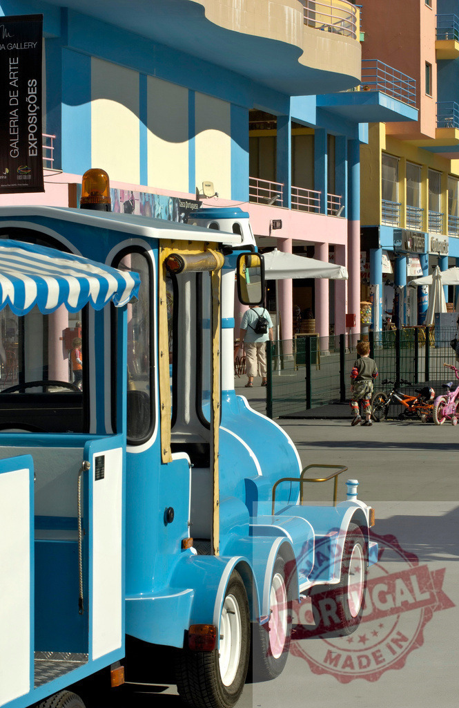 Blue tourist train on promenade near colorful buildings | Blue tourist road train parked on a promenade with vibrant buildings and people enjoying a sunny day - Realisiert mit Pictrs.com