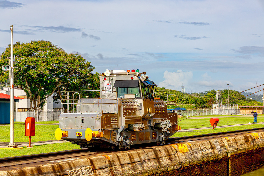 Eine Treidellokomotive am Panamakanal,„Maultier“ (Mule) | Das Bild zeigt eine spezielle Treidellokomotive am Panamakanal, die dort als „Maultier“ (Mule) bekannt ist.Zweck und Funktion -- Führung der Schiffe: Die Lokomotiven fahren auf Zählschienen an den Rändern der Schleusenkammern entlang.Seitliche Stabilisierung: Sie verbinden sich über starke Stahlseile mit dem Schiff. So halten sie den Frachter exakt in der Mitte, damit er die Betonwände nicht berührt - Realisiert mit Pictrs.com