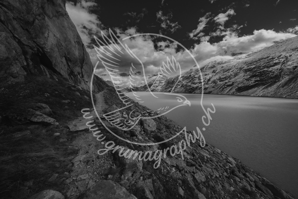 along the silent shore - switzerland_swx | Diese Aufnahme zeigt einen schmalen Weg entlang eines hochalpinen Stausees in den Schweizer Alpen. Steile Felsflanken, ruhiges Wasser und ein zurückhaltendes Wolkenbild prägen die Szene. Gedämpfte Farben, klare Flächen und eine reduzierte Bildsprache betonen die stille Atmosphäre und die Weite der Landschaft. Die Fotografie wurde bewusst ohne Inszenierung aufgenommen und im charakteristischen Grumagraphy-Style entwickelt, mit Fokus auf Ruhe, Zeitlosigkeit und natürlicher Balance.